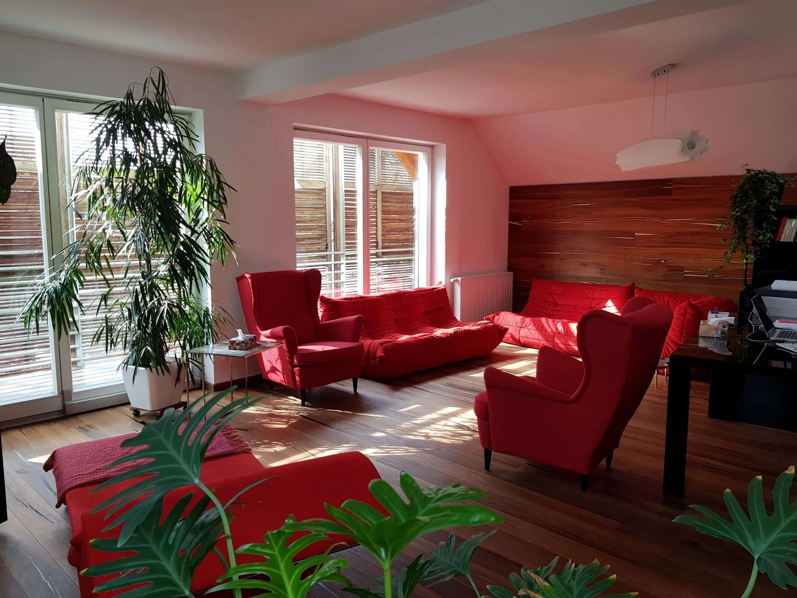 Gemütlicher, einladender Psychotherapieraum mit modernem, minimalistischem Design. Ein bequemer roter Sessel, ein stilvoller Tisch sorgen für eine ruhige Atmosphäre. Großzügige Fenster lassen viel Tageslicht herein, während warmer Holzfußboden und dezente Dekoration den Raum abrunden.