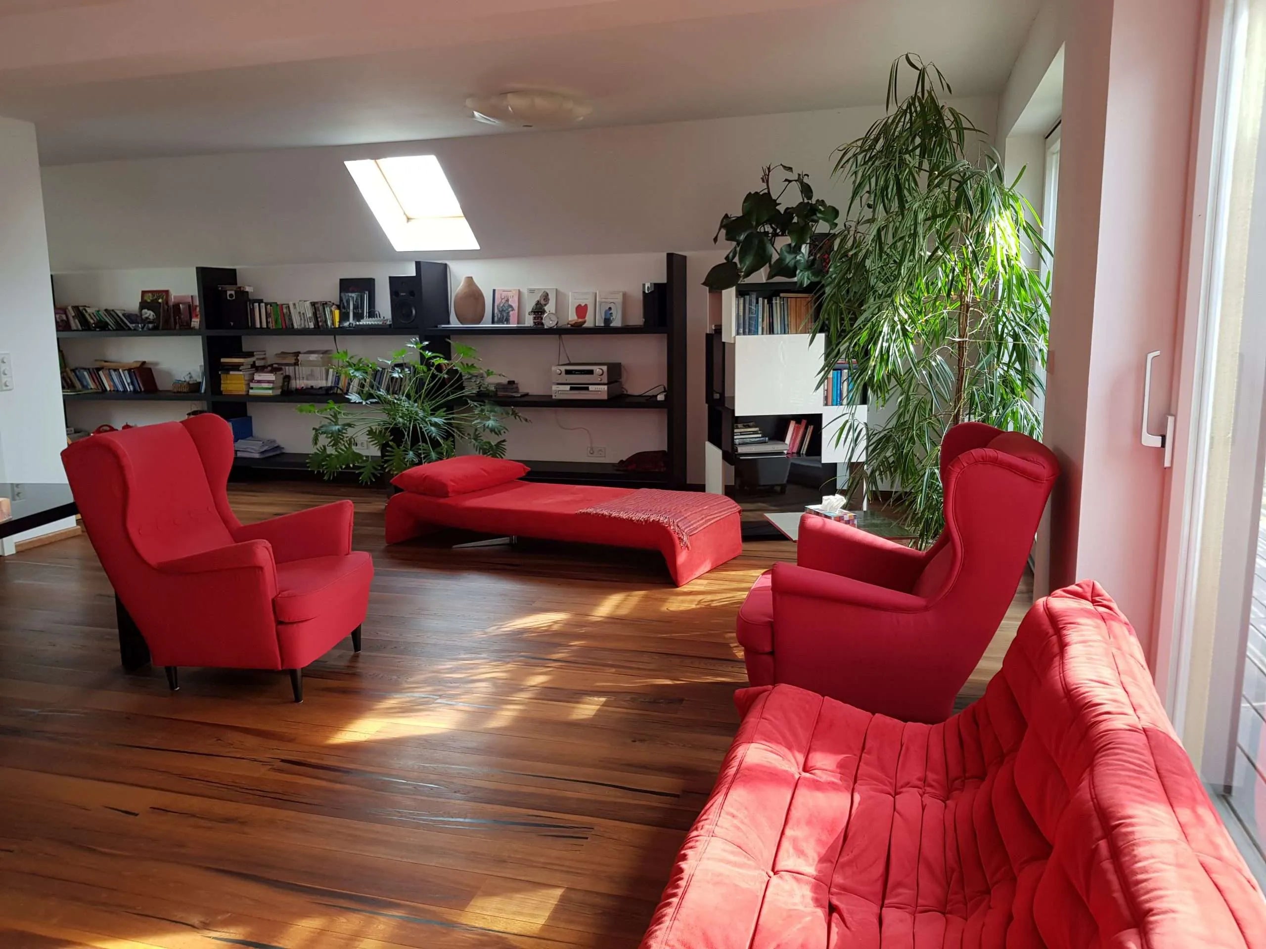 Gemütlicher, moderner Psychotherapieraum von der Seite fotografiert. Ein bequemer roter Sessel und ein stilvoller Tisch stehen vor großen Fenstern. Warmer Holzboden und dezente Dekoration unterstreichen die einladende, ruhige Atmosphäre.