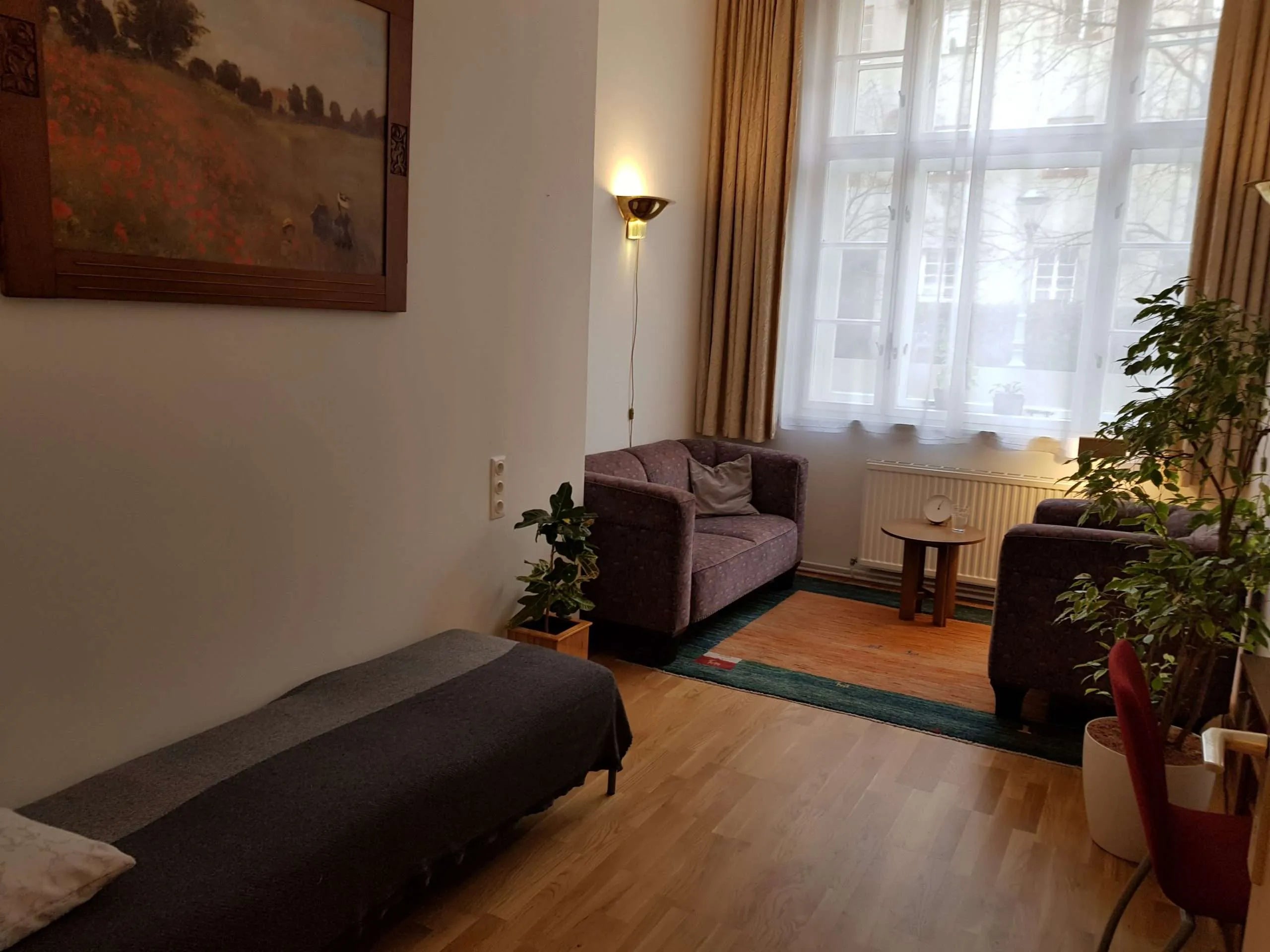 Geräumiger, lichtdurchfluteter Psychotherapieraum mit warmem Holzfußboden, bequemer Sitzgruppe und einem schlichten Couchtisch. Große Fenster lassen viel Tageslicht herein, während neutrale Farben und dezente Dekorationen eine beruhigende, einladende Atmosphäre schaffen.