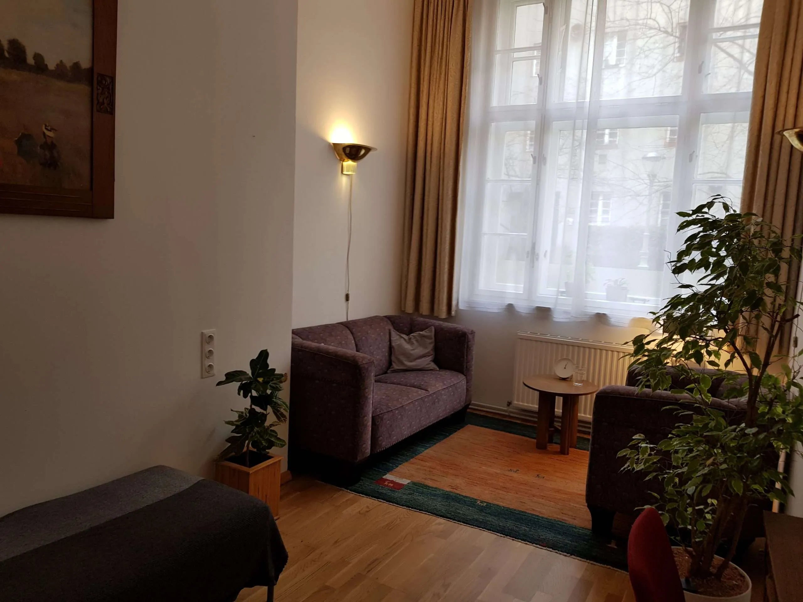 Gemütliches Therapiezimmer mit einem Sofa und passendem Sessel auf warmem Holzboden. Große Fenster lassen viel Licht herein, während neutrale Farben und dezente Dekoration für eine einladende, beruhigende Atmosphäre sorgen.